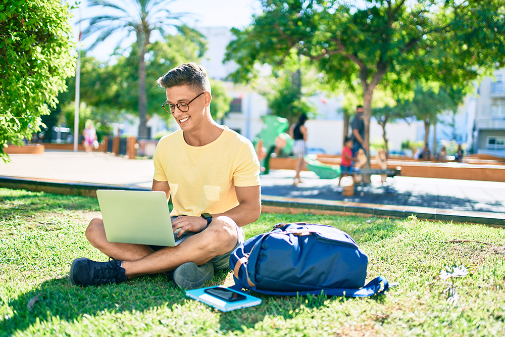 Young hispanic student smiling happy using laptop sitting on the grass at the university campus